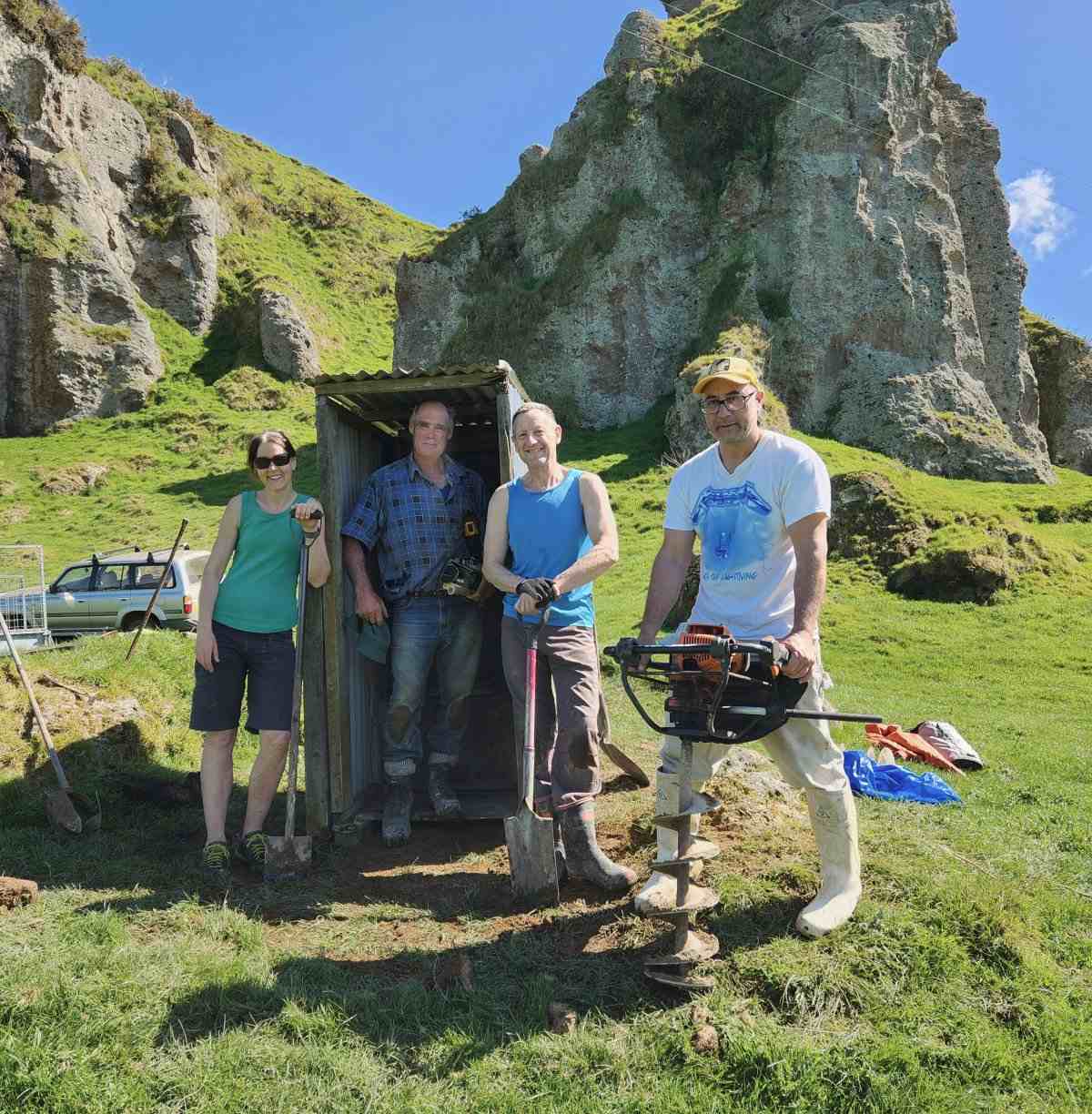 Farmer and volunteers with tools working on toilet structure with grass and cliffs in background