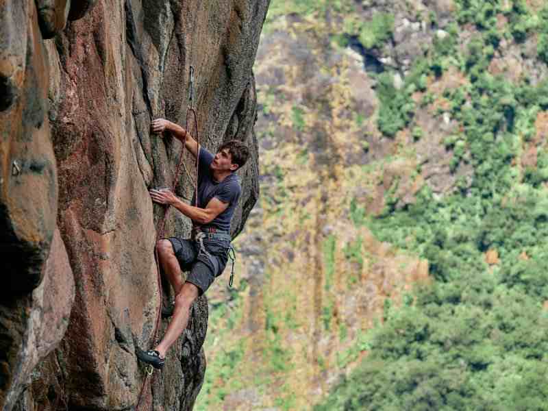 Climber wearing navy blue on sport climb. Steep orange rock with blurred trees in background.